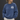 A woman wearing a washed blue crewneck sweatshirt with a white graphic print on the left chest that reads 'GOOD QUALITY HUMAN' and 'SAN ANTONIO, TX'. She is paired with off-white pants, standing against a dark gray studio background.