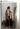 A person taking a mirror selfie in a bedroom setting, wearing a beige double-breasted blazer over a white shirt and tie, black wide-leg pants, burgundy heels, and holding a burgundy tote bag. The top of the image shows a social media caption header with the text 'kristindawnmejia Pharrell - That Girl (feat.